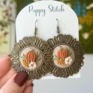 May include: A pair of handcrafted earrings featuring embroidered pumpkins, berries, and cotton on a tan background, set within ornate bronze-colored filigree frames. The earrings are displayed on a white card with the text "Poppy Stitch".
