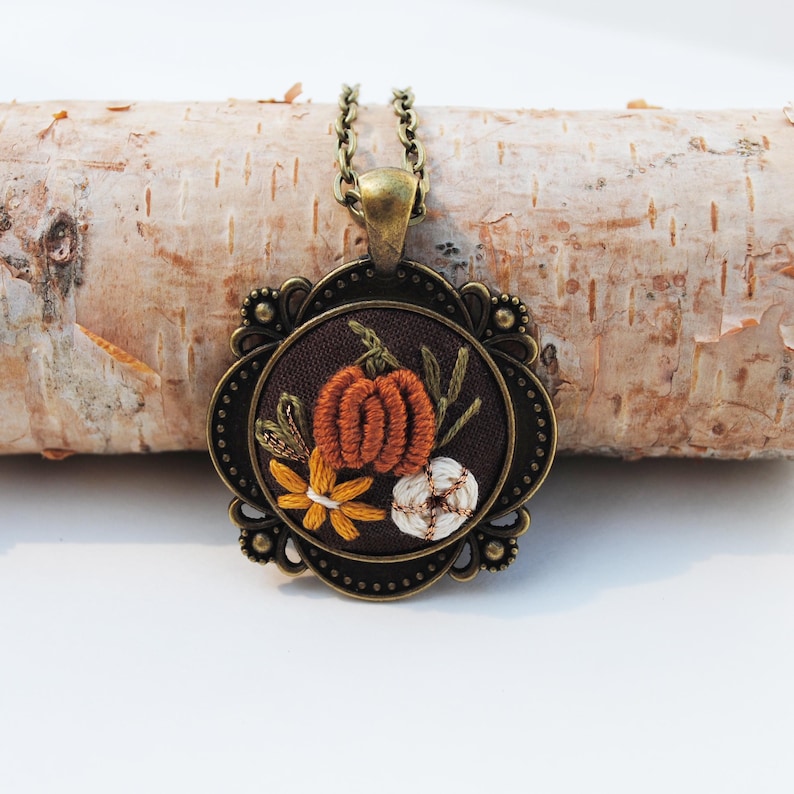 May include: A round pendant necklace featuring an antique bronze-toned metal frame with ornate detailing. The center displays dark brown fabric embroidered with a textured orange pumpkin, a small white flower, and a yellow flower with green stems.