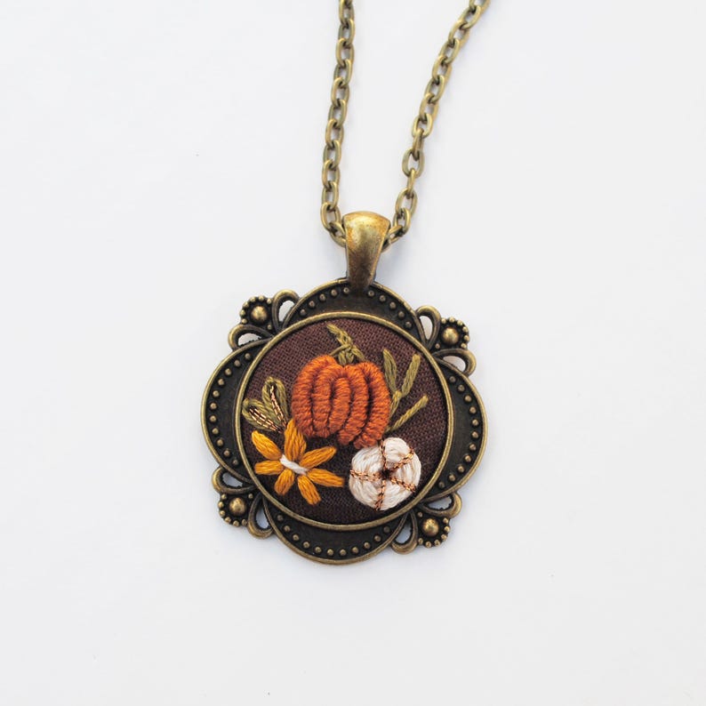 May include: A necklace featuring a round, bronze-toned metal pendant with an ornate, scalloped edge. The center contains dark brown fabric embroidered with an orange pumpkin, a white pumpkin, and a yellow flower with green stems.