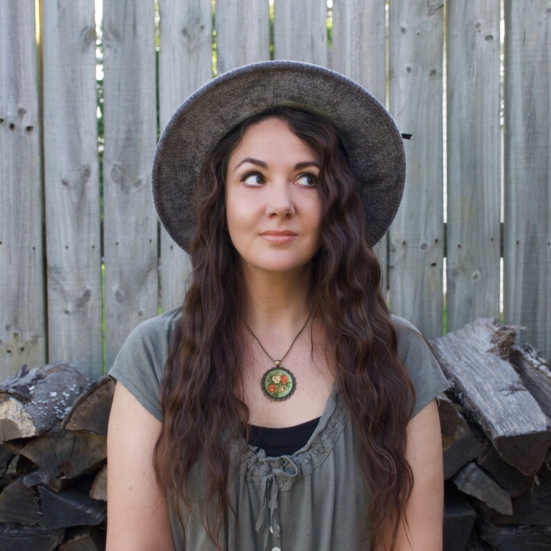 May include: A woman with long brown hair wearing a gray hat and a green dress with a floral necklace. She is standing in front of a wooden fence.