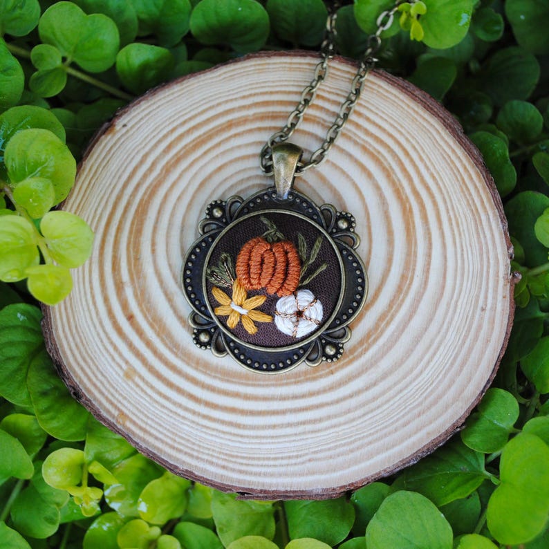 May include: A pendant necklace featuring a round, dark brown fabric insert embroidered with a central orange pumpkin, a small white pumpkin, and a yellow flower, all encased in an ornate, antique bronze-toned metal frame with a matching chain.