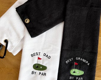 May include: Two white and black golf towels with embroidered text. The white towel says "Best Dad By Par" with a green golf course and red flag. The black towel says "Best Grandpa By Par" with a green golf course and red flag.