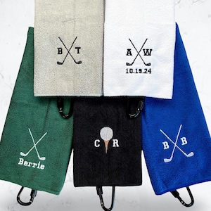 May include: Five golf towels with embroidered initials and golf club designs. The towels are in various colors, including green, blue, black, white, and gray. The towels have a loop for hanging.