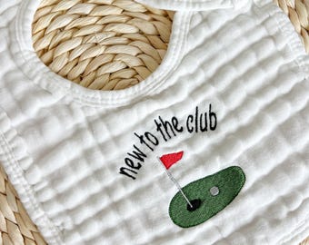 May include: White cotton baby bib with a green golf course and red flag embroidered on it. The text "new to the club" is embroidered in black.