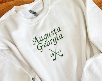 May include: White sweatshirt with green embroidered text that reads "Augusta Georgia 1932" with two crossed golf clubs.