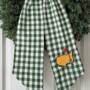 May include: A green and white gingham scarf with an embroidered golf course logo and a red flag. The scarf is draped over a green wreath. The scarf's pattern is a classic checkered design.
