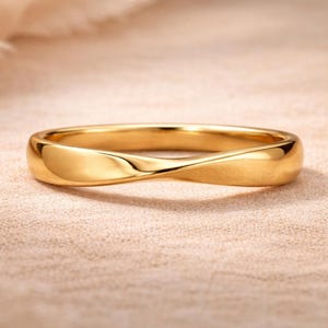 Può includere: Un anello color oro con un design a torsione. L'anello ha una superficie liscia e lucida e una forma arrotondata. L'anello è posto su uno sfondo neutro.