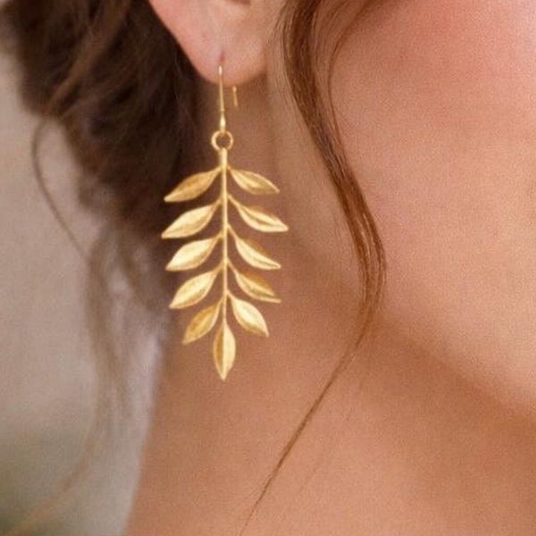May include: Gold-colored leaf earrings. Each earring features a delicate, stylized leaf design. The earrings are suspended from a small hook. The earrings are a warm gold color and appear to be made of metal. The earrings are a jewelry item.