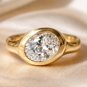 May include: A gold ring featuring an oval diamond set in a bezel. The band is smooth, polished gold. The ring is displayed on a cream satin surface. The diamond's dimensions are approximately 1.5 cm x 1 cm.