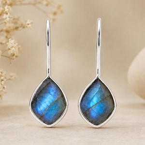 Puede incluir: Un par de pendientes colgantes de plata con piedras de labradorita en forma de lágrima. Las piedras muestran una iridiscencia azul y verde vibrante, engastadas en un marco de plata pulida. Los pendientes cuelgan de un gancho simple y elegante.