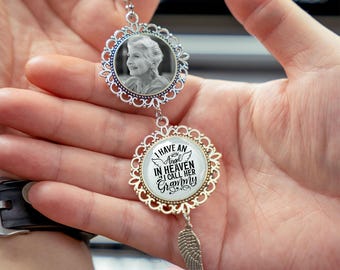 May include: A silver car charm with two circular pendants. The top pendant features a black and white photo of a woman. The bottom pendant has the text "I HAVE AN ANGEL IN HEAVEN I CALL HER Grammy". A silver feather charm hangs below.