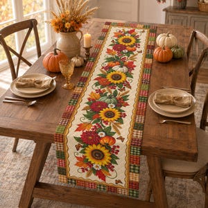 May include: A rectangular table runner featuring a central print of sunflowers, autumn leaves, pumpkins, and berries, bordered by a multicolored plaid pattern. The runner is displayed on a wooden table with place settings, pumpkins, and a vase.