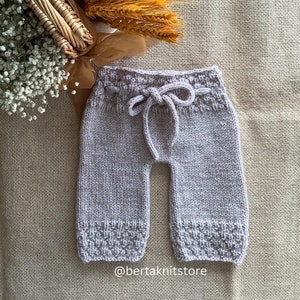May include: A pair of light grey knitted baby trousers with a drawstring waist. The trousers have a textured pattern and are made with a soft, comfortable yarn.