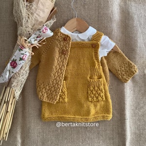 Peut inclure: Une robe bébé en tricot jaune moutarde avec un cardigan assorti et une chemise blanche à col. La robe a deux poches et le cardigan a une fermeture à bouton. La tenue est disposée sur un fond en toile de jute brune. Le texte "@bertaknitstore" est visible dans le coin inférieur droit de l'image.