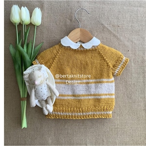May include: Mustard yellow and white striped knit baby sweater with a white Peter Pan collar, hanging on a wooden hanger. A stuffed bunny and white tulips are next to the sweater. The text "@bertaknitstore Design" is on the sweater.