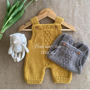Könnte beinhalten: Ein senfgelber Strickstrampler mit zentralem Zopfmuster und zwei kleinen herzförmigen Knöpfen an den Trägern, kombiniert mit einem grauen Strickstrampler mit ähnlichem Design, hängend an einem Holzkleiderbügel neben einem kleinen Plüschtier.