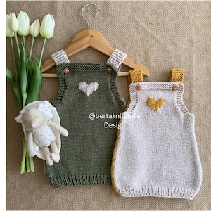 May include: Two knitted baby rompers, one olive green and one off-white, each with a heart design. The green romper has a white heart, and the off-white romper has a yellow heart. Both are on a wooden hanger. A stuffed bunny and tulips are also in the image.