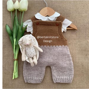 Puede incluir: Un pelele de bebé tejido en marrón y gris con una camisa blanca con cuello. Un pequeño juguete de conejito de peluche y un ramo de tulipanes blancos están junto al conjunto. El texto "@bertaknitstore Design" es visible en el pelele.