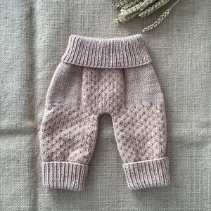 May include: Pink knitted baby trousers with a gold dot pattern. The trousers have a fold-over waistband and cuffed legs.