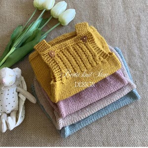 May include: A stack of three knitted dungarees in mustard yellow, dusty pink, and light blue. The top mustard yellow piece features two small heart-shaped buttons on the straps and a ribbed chest panel. Text reads Berta knit Store DESIGN.