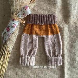 May include: Knit baby trousers in beige, orange, and brown with a ribbed texture. The trousers have a wide waistband and cuffed legs.