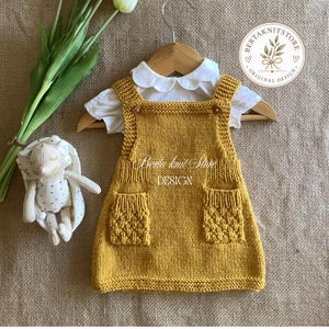 Puede incluir: Un vestido tipo pichi de punto color amarillo mostaza con dos bolsillos delanteros de patrón de rombos texturizado, sobre una camisa blanca de manga corta con cuello festoneado. Incluye pequeños botones marrones y el texto Berta Knit Store DESIGN.