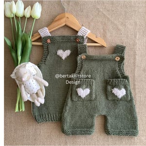 Könnte beinhalten: Zwei olivgrüne Baby-Latzhosen aus Strick mit weißen Herzdetails und Holzknöpfen. Die Latzhosen werden auf einem Holzkleiderbügel präsentiert. Ein Stoffhase und weiße Tulpen sind im Hintergrund. Der Text "@bertaknitstore Design" ist sichtbar.