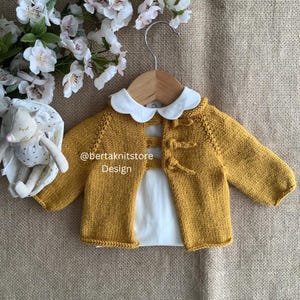 Può includere: Cardigan per neonati in maglia giallo senape con colletto Peter Pan bianco e chiusure a toggle. Il cardigan è esposto su una gruccia di legno. Il testo "@bertaknitstore Design" è visibile.