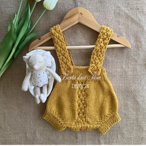 Puede incluir: Un pelele de punto color amarillo mostaza con un patrón de trenzas texturizado en el frente y los tirantes, con pequeños botones de madera en los tirantes y en la parte inferior. El texto Berta knit Store DESIGN está impreso en blanco en el centro.