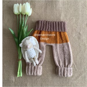 Pode incluir: Calças de bebé de malha em castanho, laranja e bege com punhos canelados. Uma boneca de coelho de pelúcia e tulipas brancas estão ao lado das calças. O texto "@bertaknitstore Design" está nas calças.