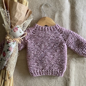 Può includere: Un morbido maglione di lana viola per un bambino o un bambino piccolo, appeso a un appendiabiti di legno su uno sfondo di juta.