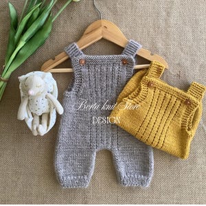 May include: A grey knitted romper and a mustard yellow knitted romper, both featuring ribbed textures and small brown buttons at the shoulder straps, displayed on a wooden hanger alongside a small white plush toy with a subtle pattern.