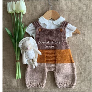 May include: A knitted romper featuring horizontal color blocks in brown, mustard yellow, and light pink, layered over a white collared shirt with a scalloped neckline. A small plush rabbit toy with gold star patterns is attached to the side.