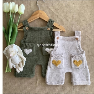 May include: Two knitted baby overalls, one olive green with a white heart pocket design, and one light gray with yellow heart pockets. The overalls are displayed on a wooden hanger. A stuffed bunny and tulips are in the background. The text "@bertaknitstore Design" is visible.