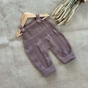 May include: A knitted baby overall in a muted mauve color, displayed on a wooden hanger. The overall features a ribbed waistband, textured panels on the legs, and button closures on the straps. The overall is made of soft yarn.