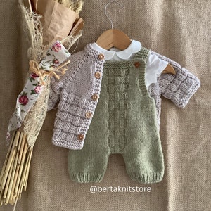 May include: A knitted baby outfit set in a light green and pale pink colour. The set includes a cardigan with buttons and a pair of dungarees. The outfit is displayed on a hanger against a hessian background.