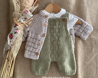 May include: A knitted baby outfit set in a light green and pale pink color. The set includes a cardigan with buttons and a pair of overalls. The outfit is displayed on a hanger against a burlap background.