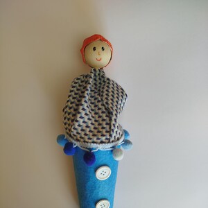 May include: A handmade doll with a wooden head, red cap, and blue and white patterned fabric. The doll has a blue felt cone body adorned with white buttons and pom-poms. A wooden stick extends from the bottom.