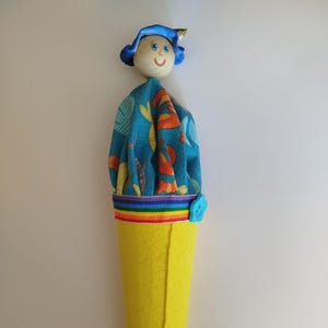 May include: A handmade doll with a wooden head, blue hat, and a smiling face. The doll's body is yellow felt, with a rainbow trim and a blue button. The doll's top is blue fabric with an orange and yellow pattern.