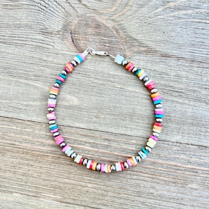May include: A colorful beaded bracelet with a silver clasp. The bracelet features a mix of vibrant, flat, round beads in shades of pink, blue, yellow, and orange, interspersed with small silver beads. The bracelet is displayed on a wooden surface.