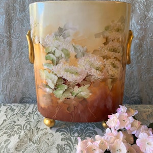 May include: A cylindrical, hand-painted vase with a floral design. The vase features a cream-colored background with a brown base and gold accents. White and green floral patterns adorn the exterior, and it has gold handles and feet. A sprig of pink flowers rests nearby.