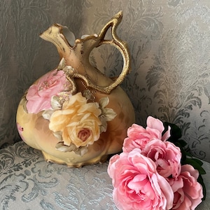 May include: A vintage, hand-painted porcelain pitcher with a gold handle and trim. The pitcher features a floral design with pink and yellow roses. A bouquet of pink roses rests beside the pitcher.
