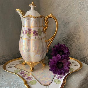 May include: A pearlescent white porcelain coffee pot with a gold handle and trim, decorated with purple flowers and green leaves. It sits on a matching tray with gold trim. A pair of purple flowers rests on the tray.