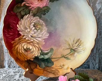 May include: A large, white porcelain plate with a gold rim features a hand-painted floral design of pink, white, and yellow chrysanthemums with green leaves. The plate is displayed on a wooden stand.