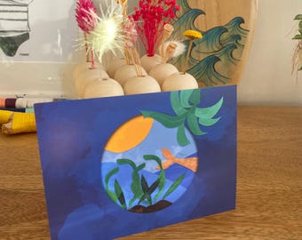 Pode incluir: Um cartão azul com um design recortado que retrata uma cena subaquática com um peixe, um sol e algas marinhas. O cartão está sobre uma superfície de madeira, com flores secas em pequenos vasos de madeira e uma prancha de surf ao fundo.