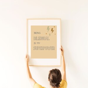 Puede incluir: Un póster beige con el texto "Ser bilingüe es mi superpoder" en texto negro y marrón con contorno. El póster está enmarcado en un marco de madera claro y se está colgando en una pared blanca.