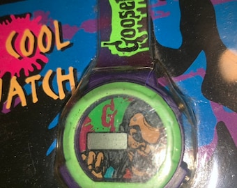 May include: A green and purple digital watch with a cartoon character on the face. The watch has a green band and the text "Goosebumps" on the side. The watch is packaged in a clear plastic case with a colorful background.