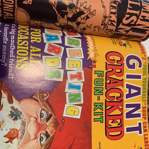 May include: A stack of vintage magazines with colorful covers. The top magazine has a peach-colored cover with black illustrations and text. The bottom magazine has a yellow cover with the text "GIANT CRACKED FUN-KIT" and a cartoon illustration.