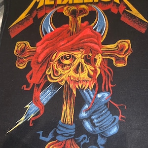 May include: Black patch with a yellow and red skull with a red bandana on its head. The skull is holding a sword and is surrounded by a yellow cross. The text "METALLICA" is in yellow letters at the top of the patch.
