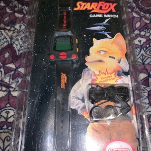 Puede incluir: Un reloj de juego Nintendo Star Fox negro con una pulsera negra y un botón amarillo. El reloj está empaquetado en una caja de plástico transparente con una imagen en blanco y negro de Star Fox en la parte posterior. El paquete también incluye unos auriculares negros.